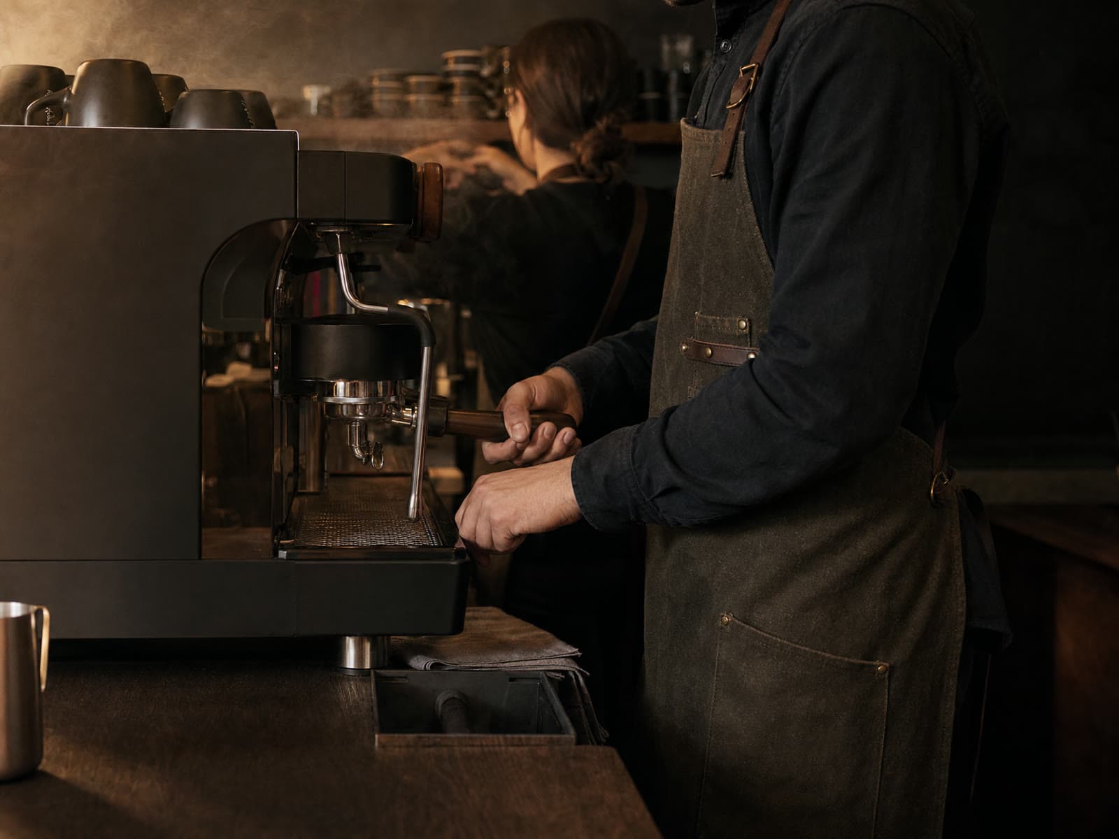 Barista fixează portafiltrul în espressor, în spatele barului; un coleg lucrează în fundal